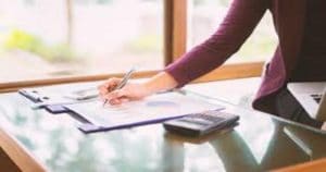 Pessoa escrevendo numa prancheta, ao seu lado, na mesa, tem uma calculadora - Conheça a tabela VRPO, que ajuda a precificar procedimentos