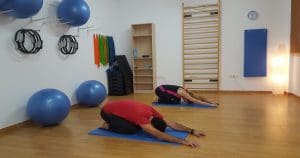 Sessão de pilates sendo realizada por um homem, que usa vermelho, e uma mulher, de roupas pretas. Ao fundo, estão instrumentos de pilates, como bolas de ginástica.