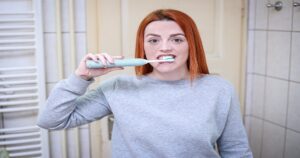 Mulher de blusa cinza em frente ao espelho escovando os dentes com uma escova de dentes elétrica azul.