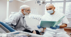 Paciente idoso sentado na cadeira do dentista enquanto o profissional mostra um caderno verde para ele.