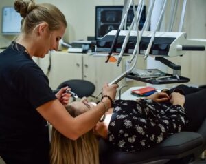 Paciente sentada na cadeira odontológica sendo atendida pela dentista. Ambas usam roupas pretas.