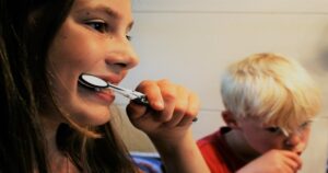 Duas crianças escovando os dentes, uma menina, que usa uma escova preta e branca, e um menino, que está de camiseta vermelha.